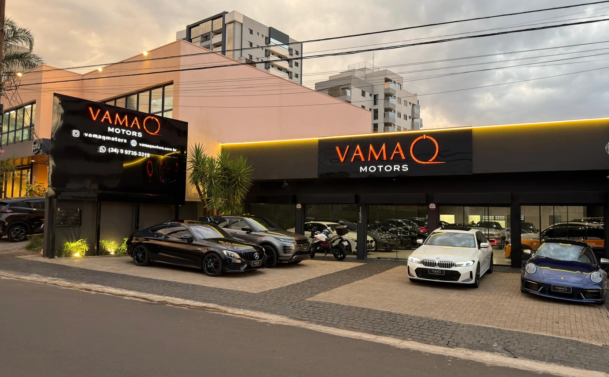 Fachada da Vamaq Motors ao anoitecer, com o letreiro aceso e carros na frente